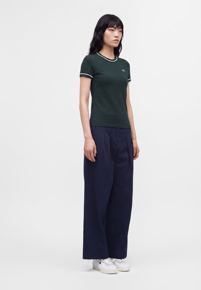 Femme portant un t-shirt ajusté vert foncé, pantalon large bleu marine et baskets blanches, debout devant un fond clair uni.