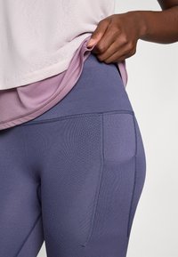 Person lifting light pink shirt to show high-waisted purple leggings with textured side pocket and stitching details.