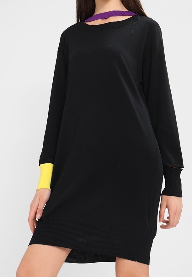 Femme portant une robe noire à manches longues avec un poignet jaune au poignet gauche et une sangle violette autour du cou sur un fond uni.
