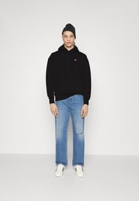 Schwarzer Hoodie mit Vorderfach und Kordelzug, kombiniert mit hellblauen, lässig destroierten Jeans. Weiße Sneakers mit schwarzen Streifen. Einfarbiger Hintergrund.