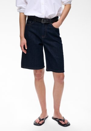 Personne portant un short en jean foncé jusqu'aux genoux, une chemise blanche à manches longues, une ceinture noire et des tongs noires, debout avec une main dans la poche.
