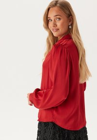 Rode, geplooide blouse met lange mouwen, voorzien van een ronde halslijn en een relaxte pasvorm. Gekocht bij een gestructureerde zwarte rok.