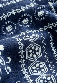 Tissu bleu avec des motifs floraux et géométriques blancs, comprenant des hexagones et des cercles, disposés en motifs décoratifs et répétitifs.