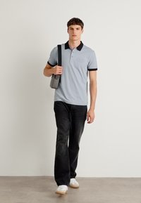 Lys blå polo skjorte med svart krage og erme aksenter, parret med mørke jeans og hvite sneakers. Modellen holder en grå skulderveske.