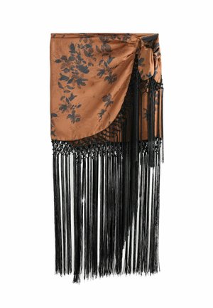 Kobieta stoi przy kamiennym murze, ma na sobie czarny top bikini i brązowy sarong z czarnymi frędzlami, obok doniczkowej rośliny na żwirowym podłożu.