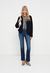 Granatowy sweter z dzianiny noszony na koszuli w kratę, połączony z rozszerzanymi niebieskimi dżinsami i jasnoszarymi szpilkami, trzymający małą kremową torebkę.