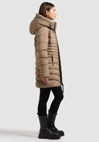Beige pufferjakke med høj krave og stor hætte, med vandret syninger, lynlåslommer og en tætsiddende silhuet. Sorte leggings og støvler.