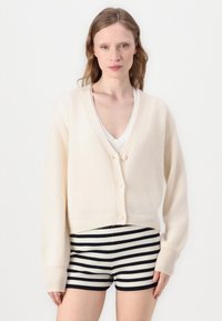 Cremefarvet cardigan med V-hals, lavet af blødt materiale, med fem knapper og ribbede manchetter. Sammen med sort-hvide stribede shorts.