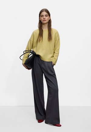 Oversized yellow sweater paired with gray wide-leg trousers. Model holds a black woven handbag with long straps and wears red heeled shoes.