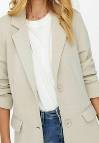 Blazer beige ligero con textura suave, dos botones y solapas con muesca. Llevado sobre una camiseta blanca y combinado con jeans azules.