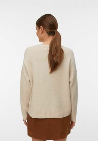 Vrouw met een lange bruine paardenstaart, gekleed in een beige gebreide trui en een bruine rok, van achteren gericht tegen een effen witte achtergrond.
