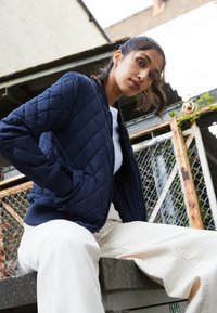 Veste bomber matelassée bleu marine avec des poignets et un ourlet côtelés, superposée à une chemise blanche, associée à un pantalon de couleur claire. Tissu texturé, design décontracté.