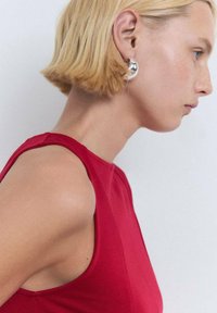 Red sleeveless top with a smooth texture. Model with short blonde hair wears round metallic earrings, showcasing a side profile.