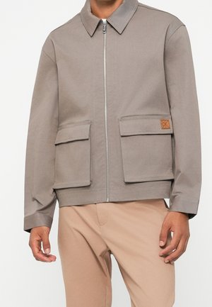 Man wearing taupe zip-up jacket with flapped front pockets and beige pants, standing against a plain background.