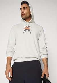 Mand iført en hvid hættetrøje med "X FIT" logo, sorte bukser, ser til siden mens han holder en sort kasket i sin højre hånd.
