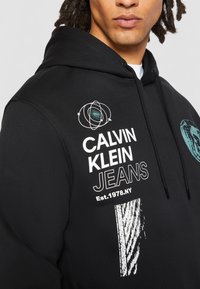 Czarna bawełniana bluza z kapturem z białymi i teowymi grafikami, w tym napisem "CALVIN KLEIN JEANS", motywem planety oraz szczegółami w paski.
