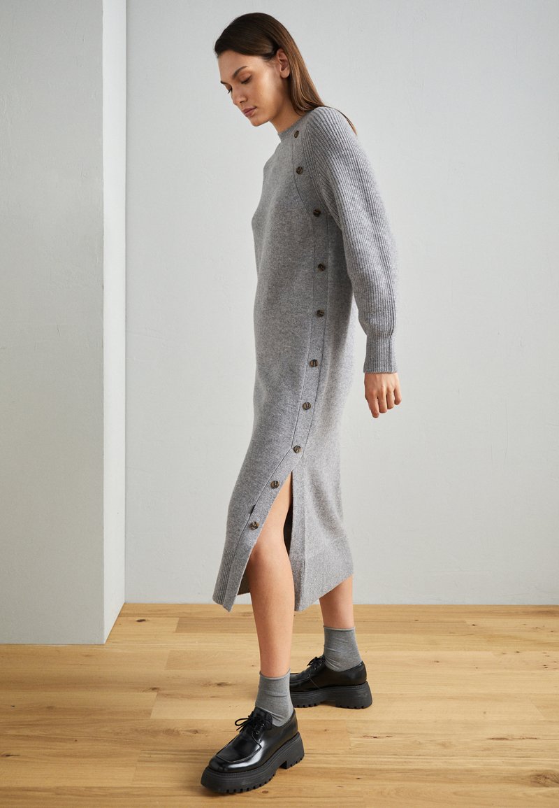 Szara dzianinowa sukienka z prążkowanymi rękawami, bocznymi guzikami i rozcięciem do uda. Stylizowana z czarnymi butami i szarymi skarpetkami na drewnianej podłodze.