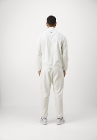 Fato de treino branco com padrão xadrez, com punhos e cós elásticos, apresentando um pequeno logótipo no pescoço. Usado com tênis de cor clara.