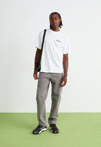 Hvit tshirt med korte ermer og "Dickies"-logo, grå, løstsittende jeans og svarte joggesko med hvite detaljer. Står på grønt gulv.
