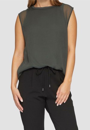 Bezrekawnik o ciemnozielonym kolorze z przezroczystymi panelami na ramionach zestawiony z czarnymi spodniami na sznurek na stojącej osobie na jednolitym tle.