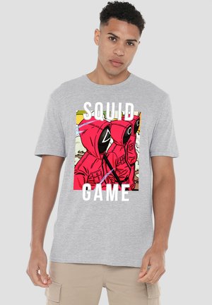 Young man wearing a gray t-shirt featuring a graphic of masked figures in red hoods with the text “SQUID GAME” below.