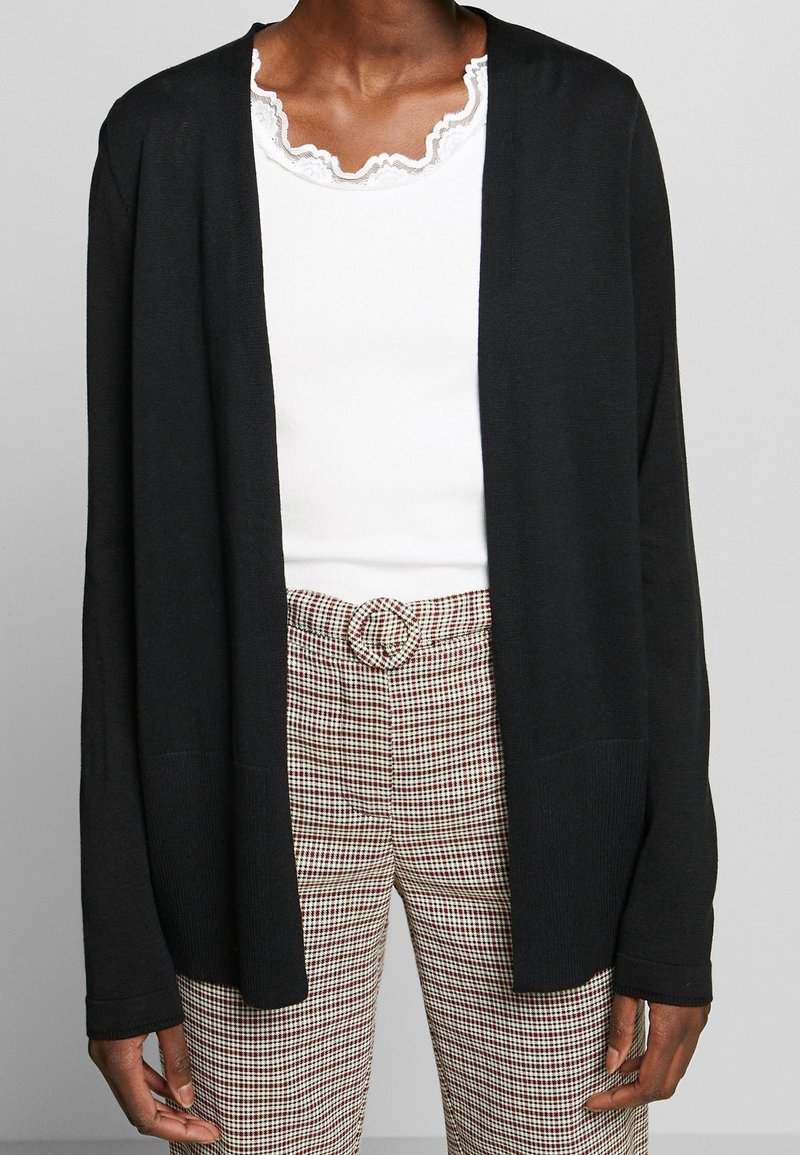 Personne portant un cardigan noir ouvert sur le devant, un haut blanc à bords volantés, et un pantalon taille haute à carreaux avec une boucle de ceinture circulaire.
