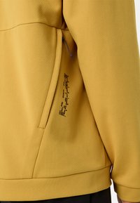 Gelbe Jacke mit Textur, die eine Reißverschlusstasche und einen kontrastierenden Kordelzug aufweist, sowie gedruckte Details an der Seite.