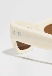 Lunettes de soleil blanches avec une monture épaisse, des verres marron et des accents texturés. Les branches présentent cinq petits points gris pour le détail.