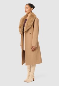 Lång kappa i camel med krage av konstpäls, dubbelknäppta knappar och rak silhuett, i kombination med långa nudefärgade stövlar i mocka.