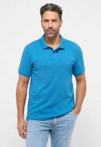 Tyrkysové polo tričko s krátkými rukávy a límečkem. Vyrobeno z hladké látky, obsahuje decentní logo na levé straně hrudi. Nosené s světlými modrými džíny.