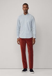 Camisa de botones a rayas azul y blanca con un bolsillo, combinada con pantalones de pana color óxido y zapatillas negras.