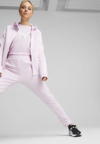 Svetlo roza športna obleka, ki vključuje zadrgo jakno in usklajene hlače, v kombinaciji z črnimi supergami. Gladka tekstura z minimalnimi oblikovalskimi detajli.