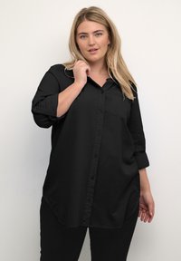 Camicia nera con bottoni, maniche lunghe, taschino sul petto, orlo curvo e texture morbida. Indossata con pantaloni neri.