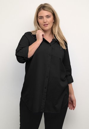 Camicia nera con bottoni, maniche lunghe, taschino sul petto, orlo curvo e texture morbida. Indossata con pantaloni neri.