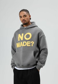 Grijze hoodie met grote gele letters "NO MADE?" op de voorkant, kangoeroezak, gestructureerde stof en een comfortabele pasvorm.