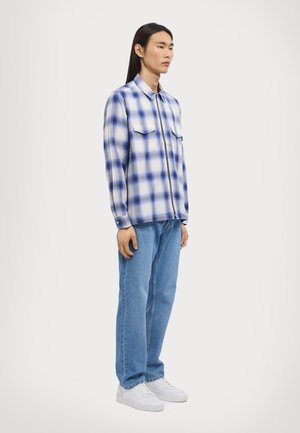 Camicia con collo in fantasia a quadri blu e bianchi, dotata di due tasche sul petto; abbinata a jeans azzurri chiari e sneakers bianche.