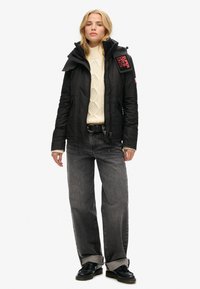 Sort isoleret jakke med aftagelig hætte, lynlås foran, der har et rødt logo på skulderen, båret over en cremefarvet kabelstrikket sweater og grå jeans.