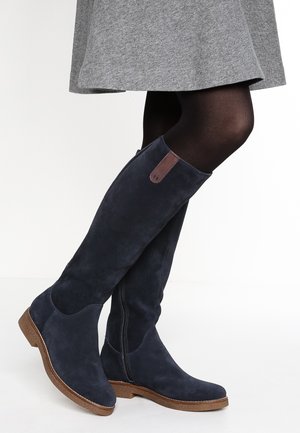 Vrouw die marineblauwe suède kniehoge laarzen draagt met een bruine zool en rits, gecombineerd met een zwarte panty en een grijze rok.