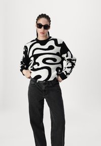 Zwart-witte abstracte gebreide trui met een ontspannen fit, gecombineerd met hoog-gesneden donkere jeans. Zwarte zonnebril maakt de look compleet.