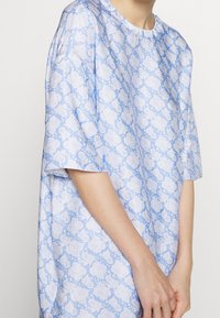 Blusa oversize azzurro chiaro con maniche corte, caratterizzata da un motivo floreale bianco e dettagli a pois su un tessuto liscio.
