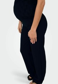 Personne enceinte portant un pantalon de maternité noir texturé et des sandales noires, debout avec une main reposant sur sa cuisse.