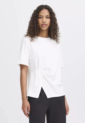 Femme aux cheveux bouclés portant un haut blanc à manches courtes avec une fente latérale et un pantalon noir, debout devant un fond blanc uni.