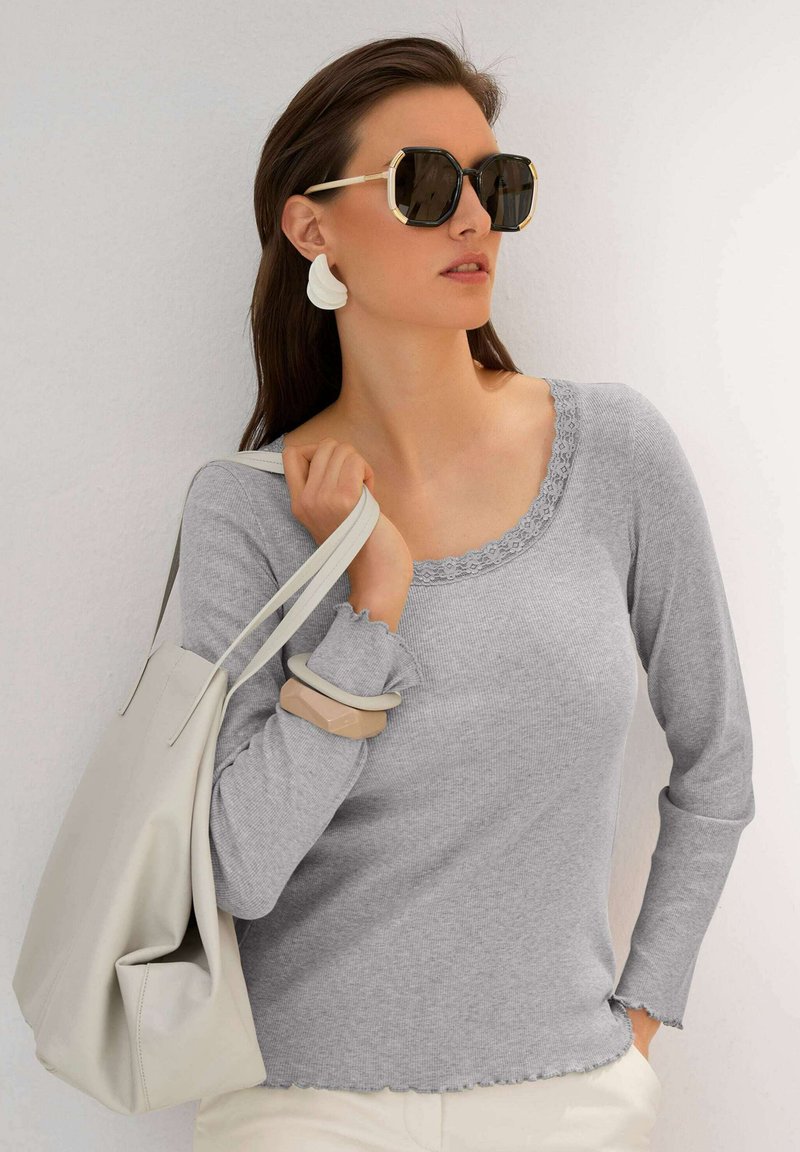 Grijze lange mouwen top met een gegolfde halslijn en manchetten, gecombineerd met een lichtgrijze tas. Het model draagt oversized zwarte zonnebrillen en witte oorbellen.