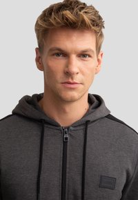 Szara bluza z kapturem na zamek błyskawiczny z czarnymi akcentami, wykonana z teksturowanej tkaniny, z sznurkiem ściągającym i prostokątną naszywką logo na klatce piersiowej.