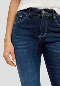 Jeans en denim bleu foncé avec une coupe ajustée, dotés de quincaillerie en argent, de cinq poches et de surpiqûres visibles.