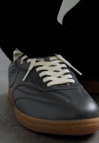 Szürke bőrsneaker sima textúrával, fehér fűzőkkel és barna gumitalppal, finom varrással és minimalistába illeszkedő dizájnnal.