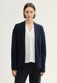 Cardigan bleu marine à ouverture devant, en tissu texturé, avec des manches longues et une coupe décontractée, porté par-dessus une blouse blanche et un pantalon sombre.
