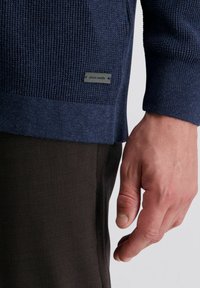 Marineblauer Strickpullover mit strukturiertem Muster am Saum, ausgestattet mit einem kleinen schwarzen Etikett. Kombiniert mit dunkelbraunen Strukturhosen.