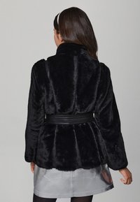 Jachetă neagră din blană artificială cu mâneci lungi și guler larg, cu curea în talie dintr-o bandă texturată neagră. Fustă metalică netedă și lucioasă.