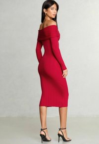 Abito rosso a coste senza spalline con maniche lunghe, silhouette aderente e lunghezza a metà polpaccio. Indossato con tacchi a spillo neri.